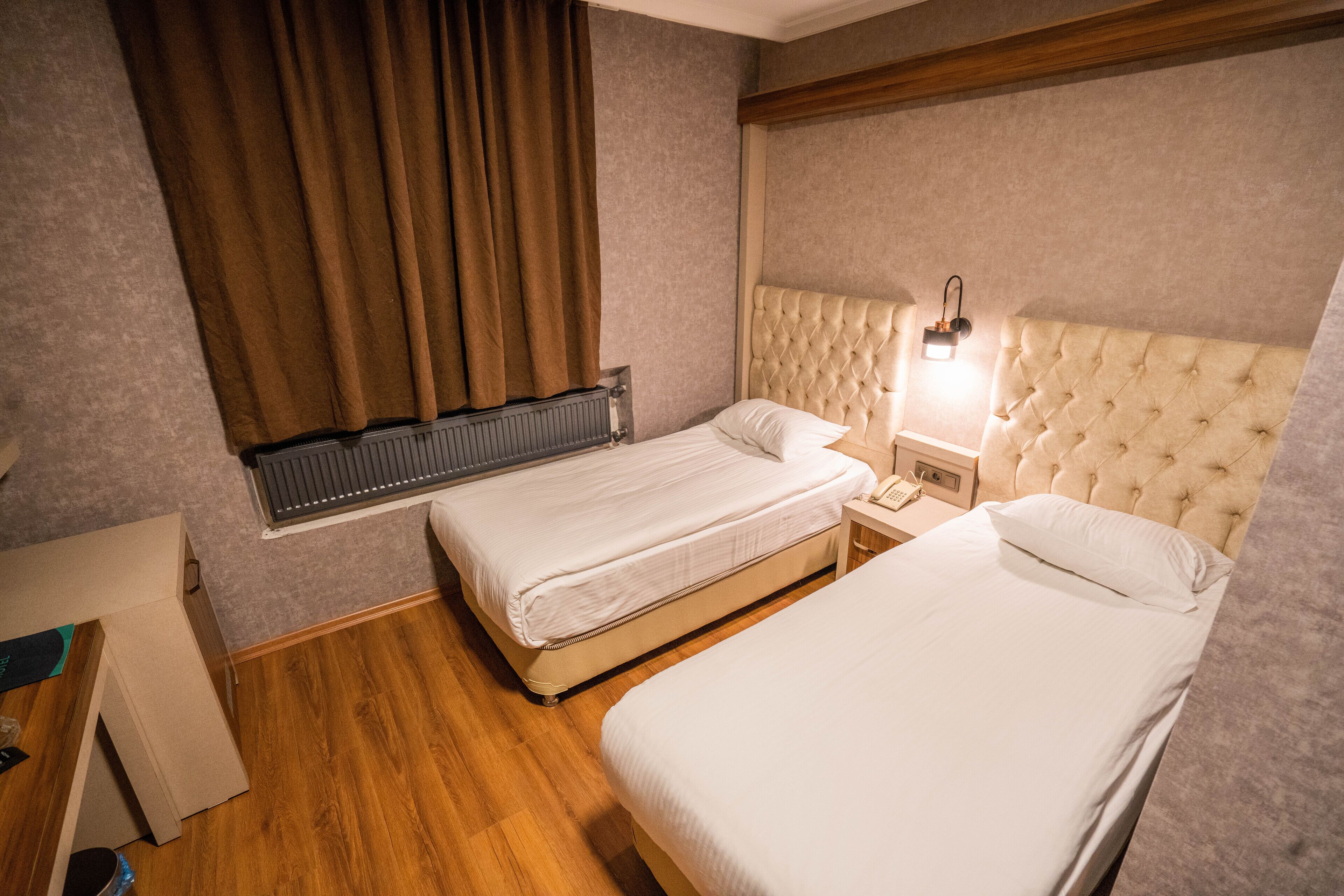 A hotel room with two single beds, a wooden floor, a radiator, a nightstand with a lamp, and a window with curtains.