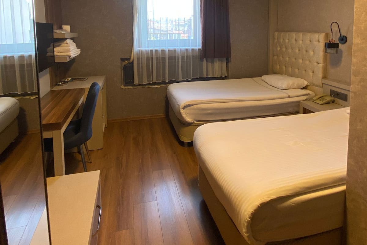 A hotel room with two beds, a desk, a chair, a bedside table with a lamp, and a window with curtains.