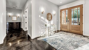 Un pasillo con piso oscuro, paredes blancas y una puerta grande de madera con paneles de vidrio. Hay apliques de pared y un espejo en la pared.