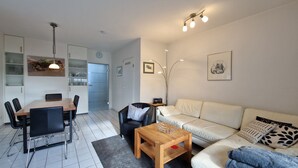 Ein modernes Wohnzimmer mit einem Holzesstisch, schwarzen Stühlen, einem weißen Sofa und einem Couchtisch. Es gibt eine Küchenzeile mit Kühlschrank, Mikrowelle und Spüle.