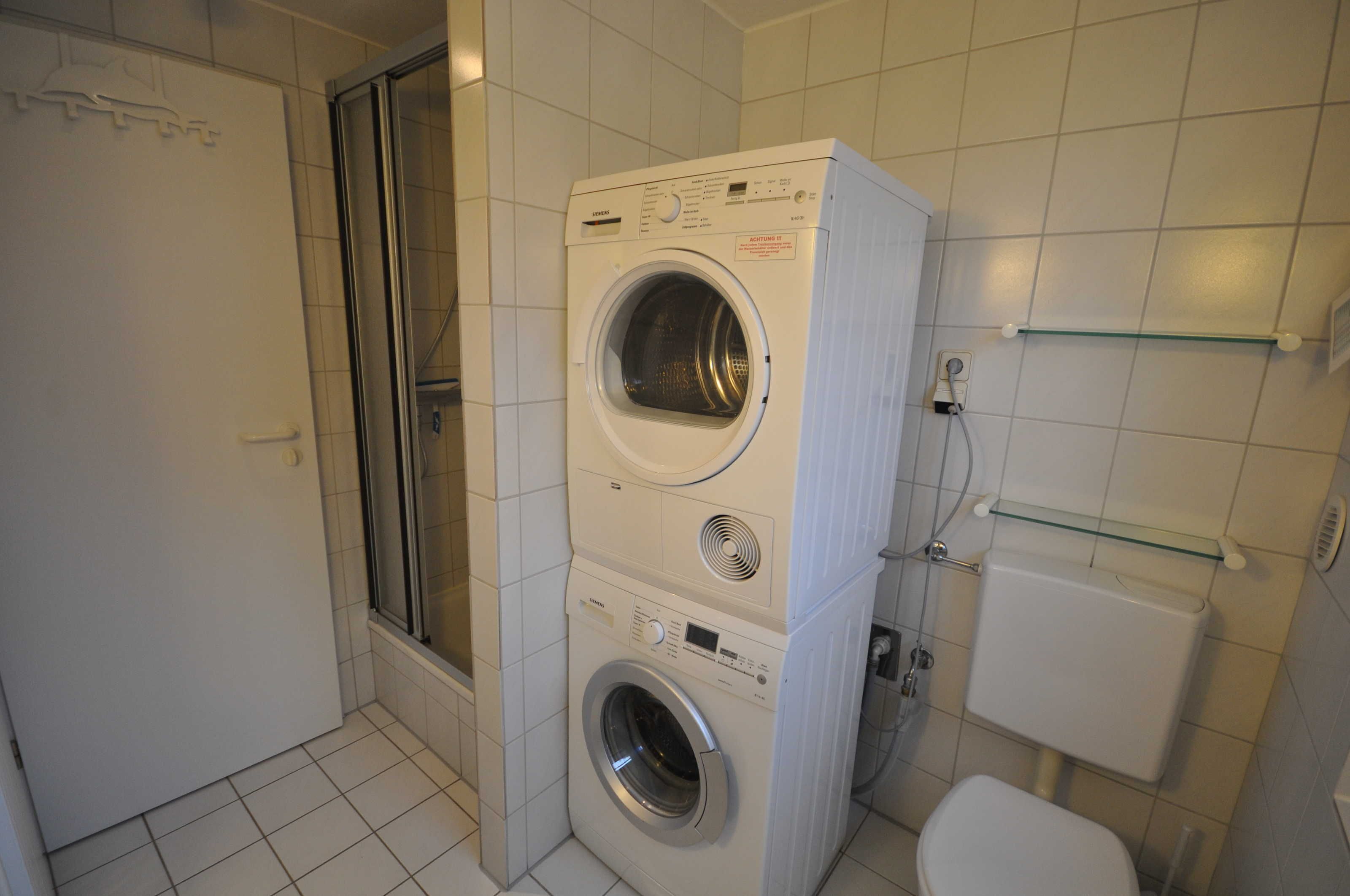 Une petite buanderie équipée d’un lave-linge et d’un sèche-linge à chargement frontal, d’une cabine de douche en verre et de toilettes.