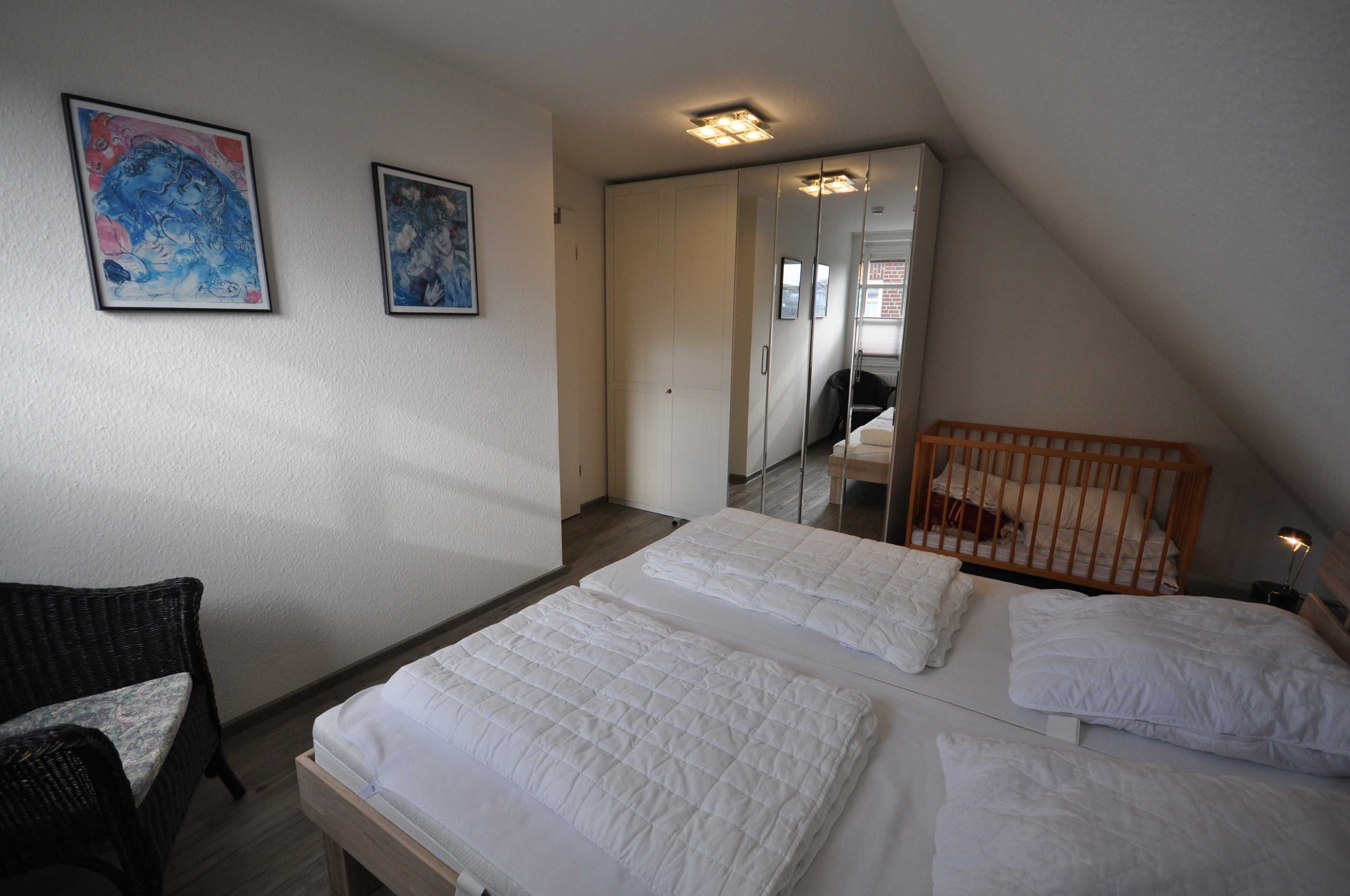 Une chambre avec deux lits, un berceau, une chaise et deux œuvres d’art encadrées accrochées au mur.