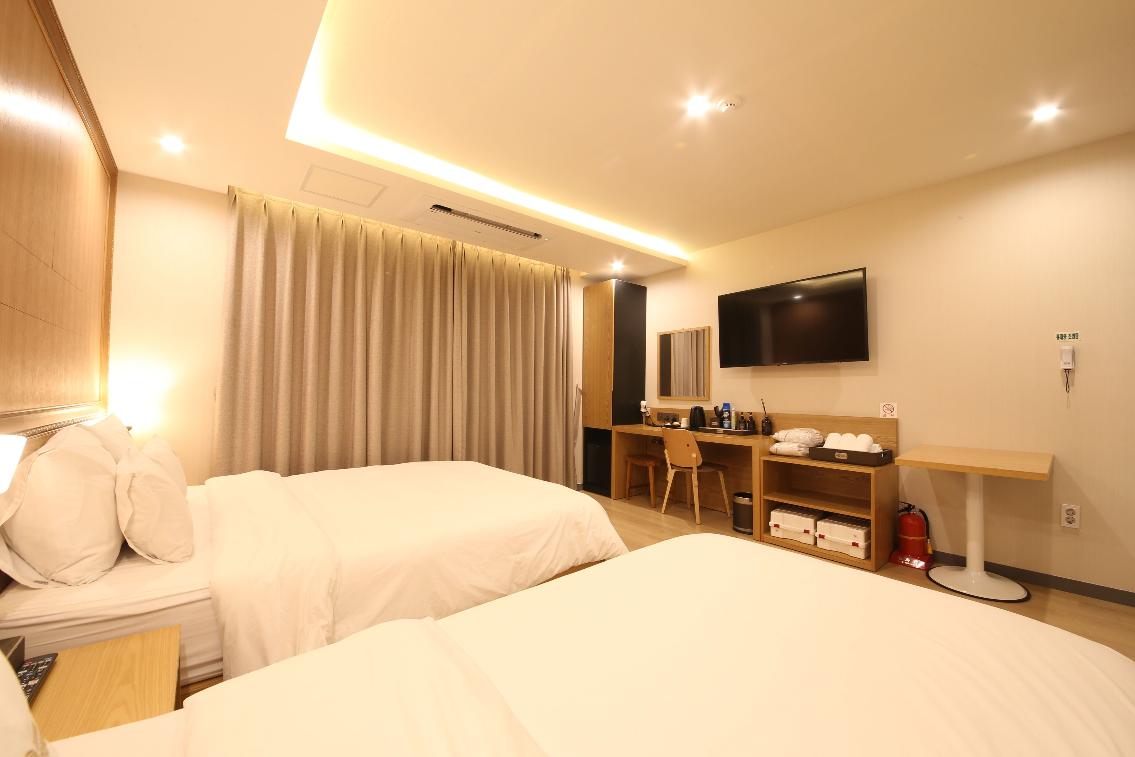 A hotel room with two beds, a desk, a chair, a television, and a fire extinguisher.