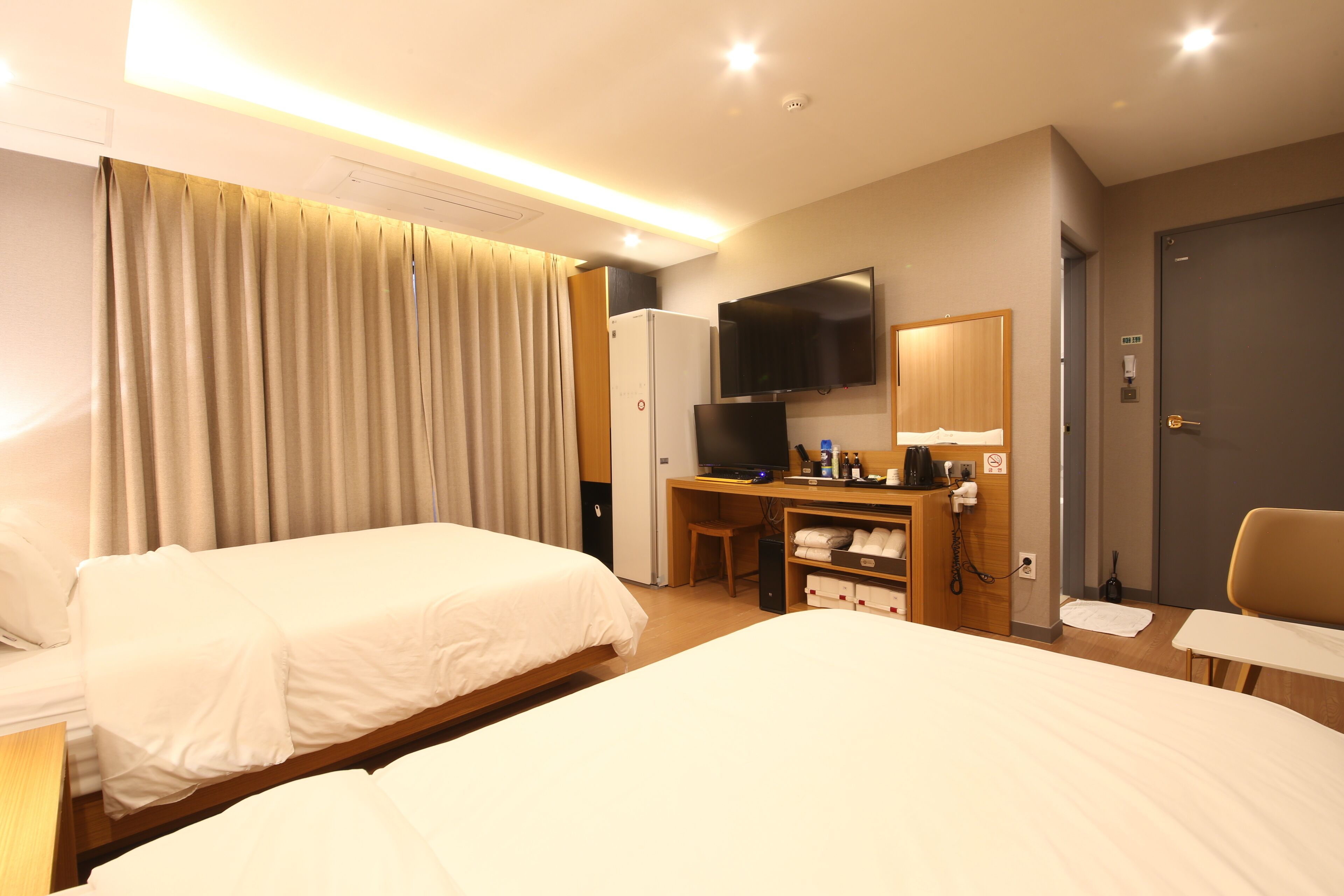 A hotel room with two beds, a desk with a TV, a minibar, and a door.