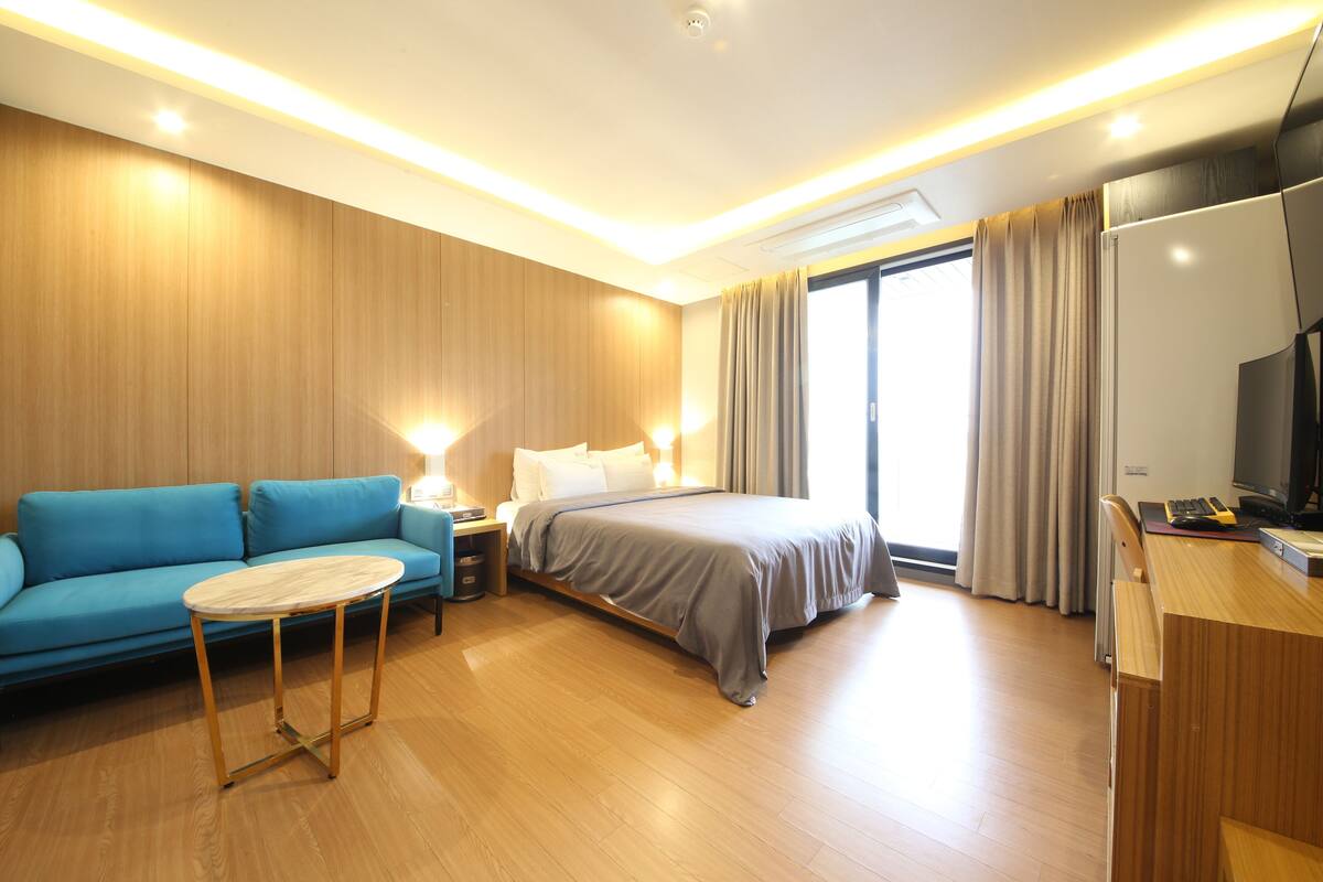 Moderni hotellihuone, jossa on sänky, sininen sohva, pieni pöytä ja työpöytä, jonka päällä on televisio.