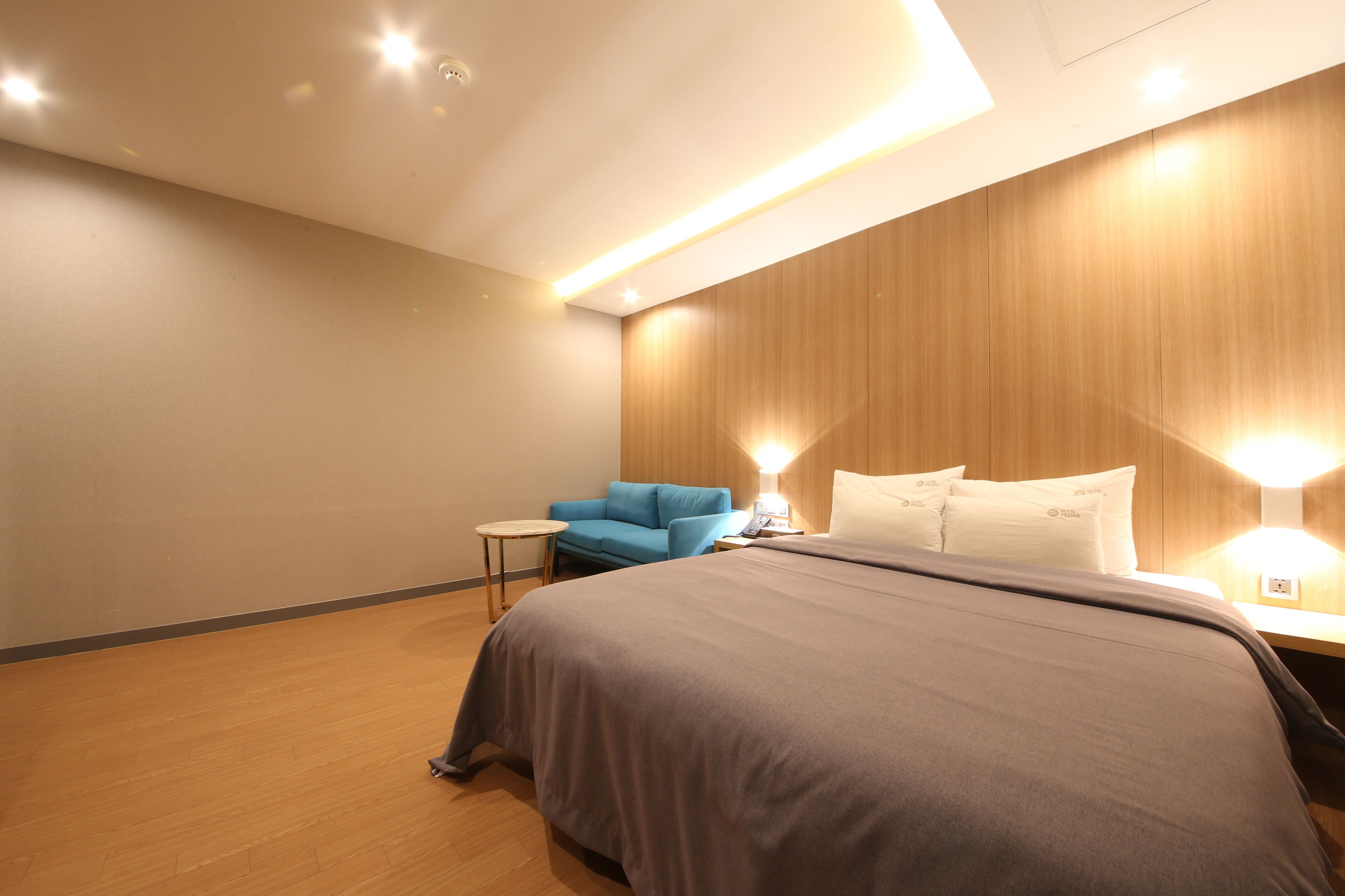 A modern hotel room with a large bed, a small side table, a blue chair, and wooden paneling on the walls.
