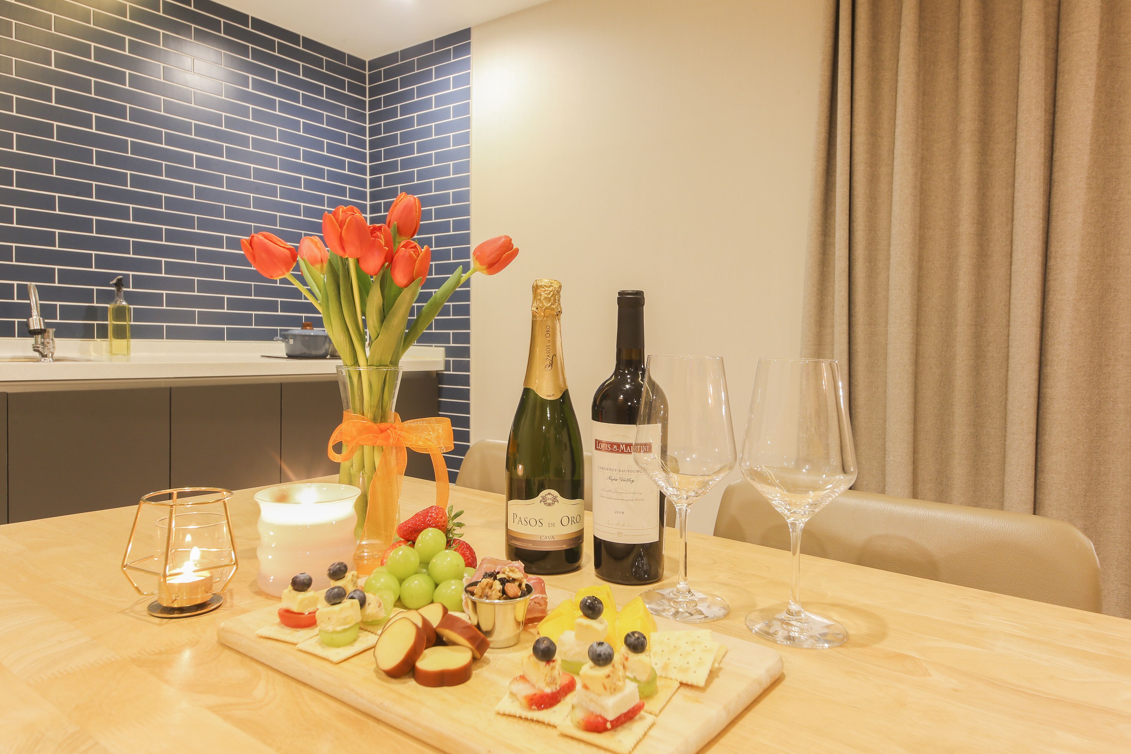 A dining table with a variety of snacks, two bottles of wine, a candle, and a vase with red tulips.