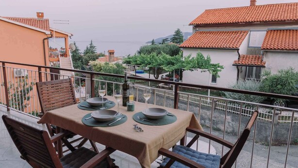 Un balcon avec une table dressée pour le dîner, donnant sur un quartier résidentiel aux toits recouverts de tuiles en terre cuite.