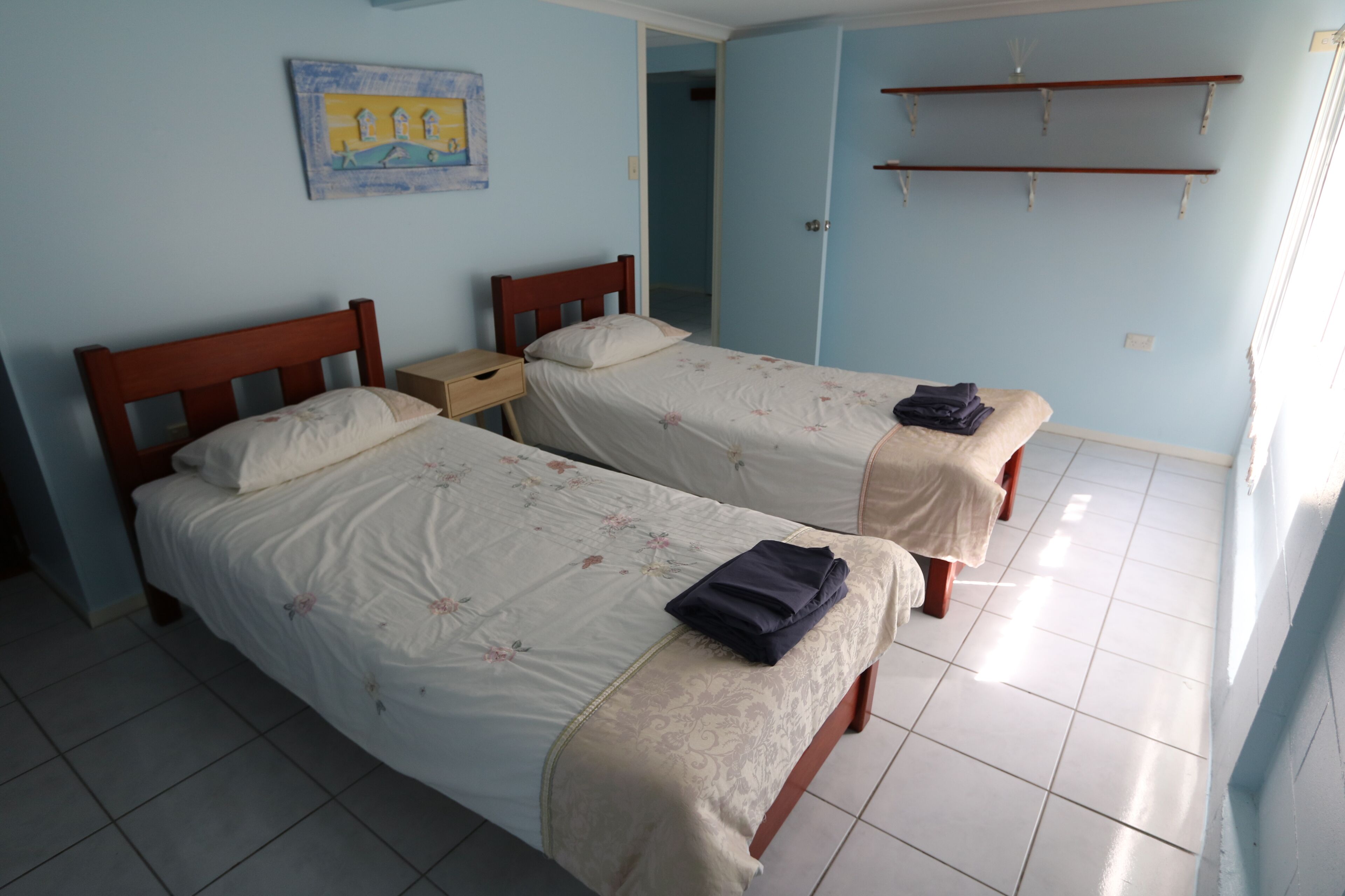 A room with two single beds, a small bedside table, a wall-mounted shelf, and a door leading to another room.