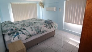 A bedroom with a bed, a standing fan, a window with blinds, and a wall-mounted air conditioner.