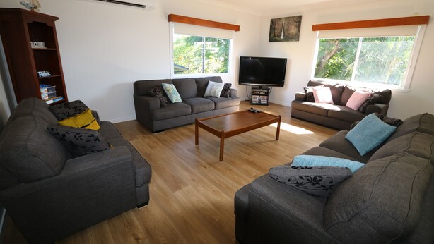 A living room with two sofas, a coffee table, and a TV. There are windows with blinds, a bookshelf, and a ceiling fan.