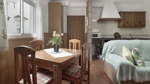 Uma sala de jantar com mesa e cadeiras de madeira, uma planta em vaso e vista para a cozinha.