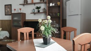 Uma sala de jantar com mesa, cadeiras, um vaso com flores, televisão e prateleiras com diversos objetos.