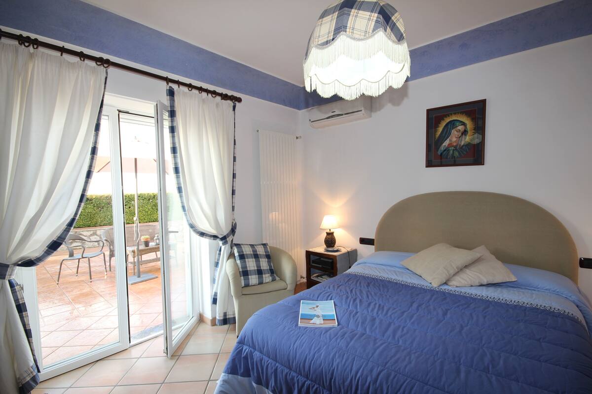 A bedroom with a bed, a nightstand, a lamp, a chair, and a view of an outdoor patio.