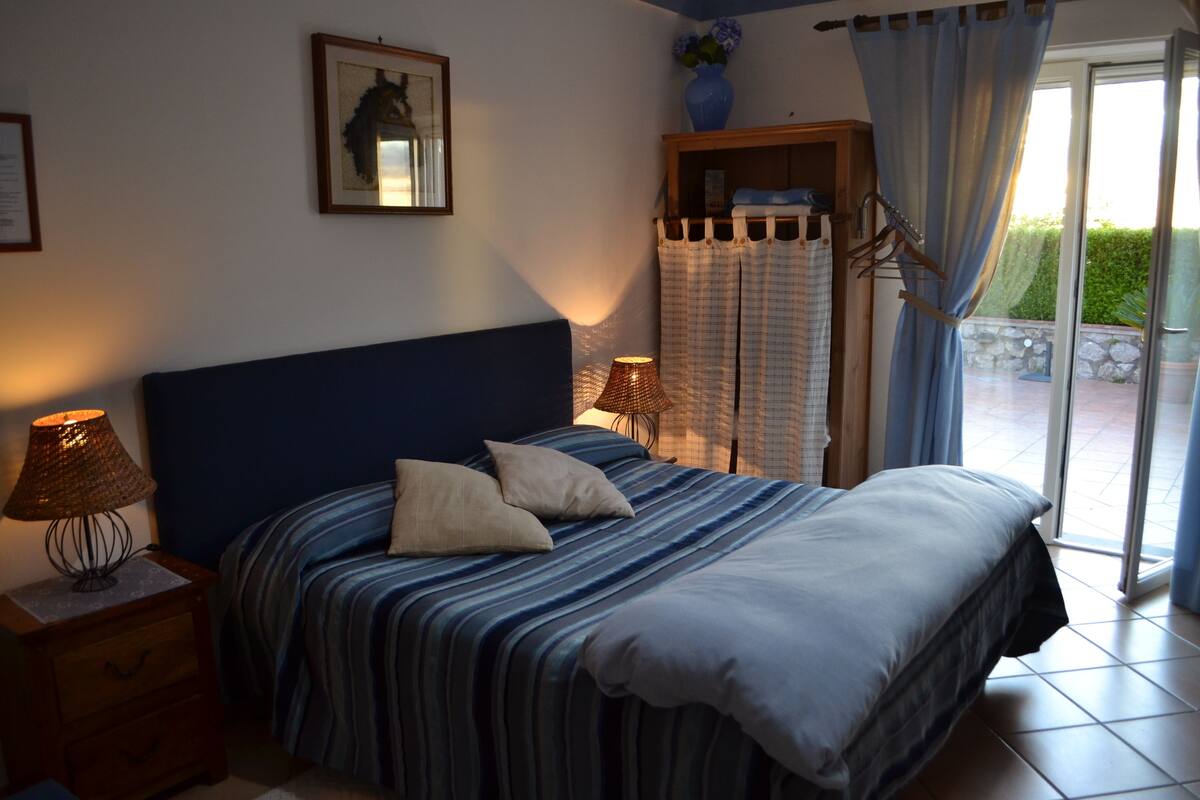 A bedroom with a bed, a nightstand, a lamp, a framed picture on the wall, and a window with curtains.