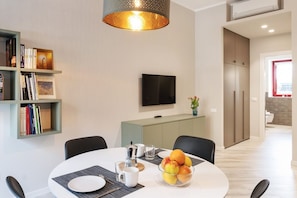 Un espace repas moderne avec une table ronde, des chaises, un saladier de fruits, une cafetière et une bibliothèque remplie de livres.
