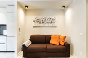 Ein Wohnzimmer mit einem braunen Sofa, zwei orangefarbenen Kissen, einer grauen Decke und einem an der Wand befestigten abstrakten Kunstwerk.