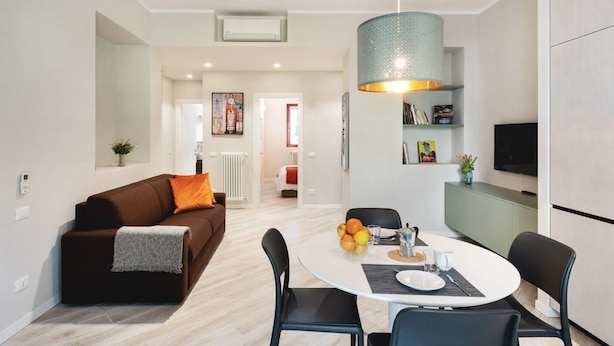 Een moderne woonkamer met een bruine bank, een eettafel met stoelen, een keukenhoek met een televisie, en een gang die naar een andere kamer leidt.