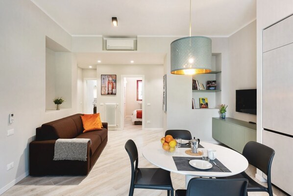 A modern living room with a brown sofa, a dining table with chairs, a kitchen area with a TV, and a hallway leading to another room.