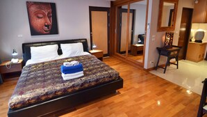 Et hotelværelse med en stor seng, trægulv, en Buddha-statue ophængt på væggen, et spejl og et lille bord med en lampe.