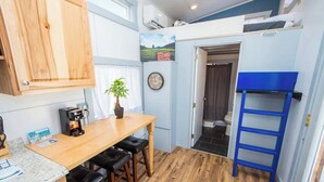 Een kleine keuken met een houten tafel, zwarte krukken, een koffieapparaat en een klok aan de muur.