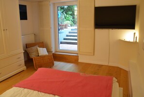 Une chambre à coucher comprenant un lit, une chaise, une commode, un téléviseur et une porte donnant sur un balcon agrémenté de plantes.