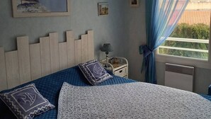 A bedroom with a bed, a nightstand, a lamp, a window with curtains, and a painting on the wall. - Central Arcachon: 400m from Beach, T2 with Balcony, Parking, and Pet-Friendly (Arcachon)