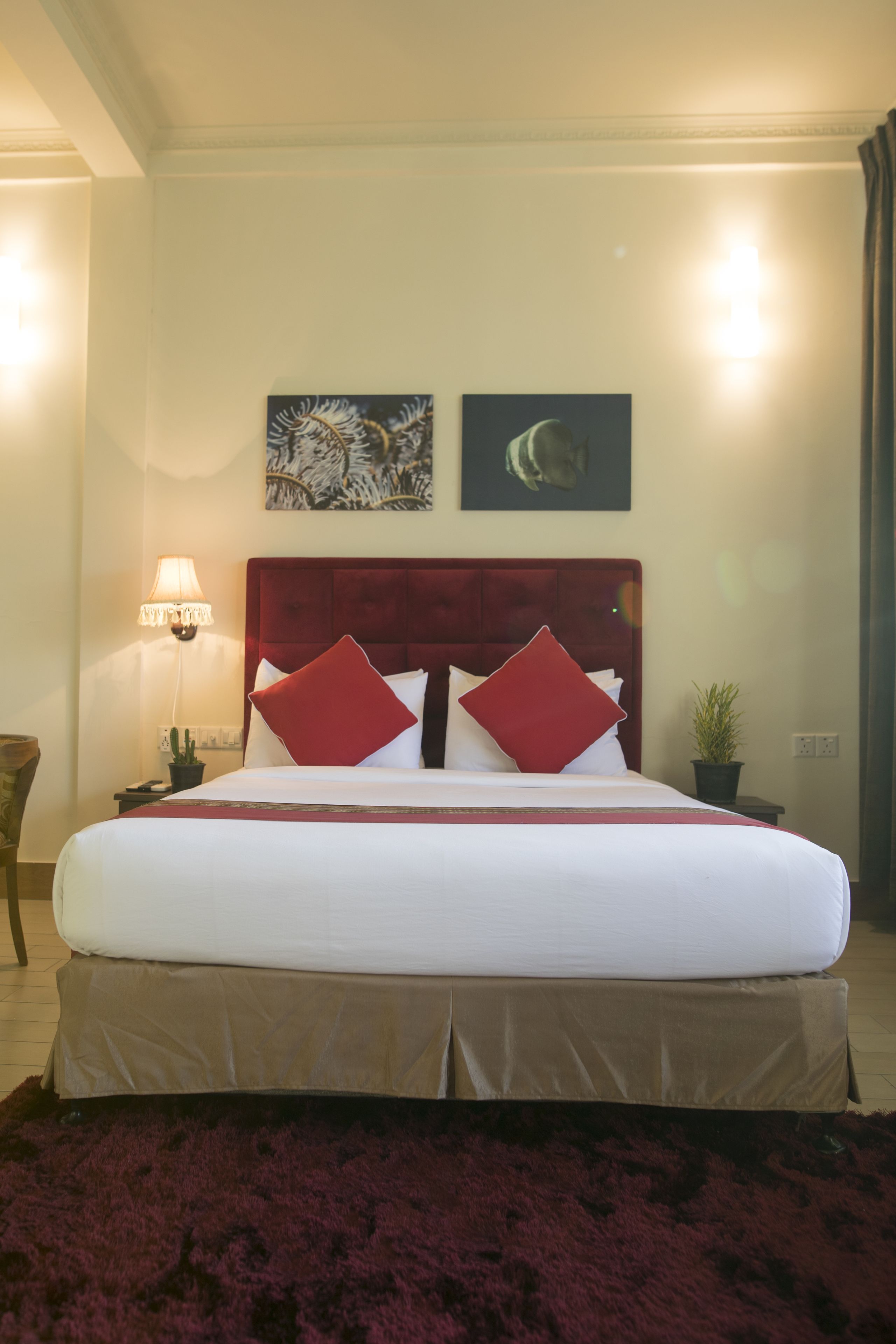 Een net opgemaakt bed met een rood hoofdeinde, witte beddengoed en rode kussens. Naast het bed staat een klein tafeltje met een lamp, en op de vloer staat een pot met een plant.