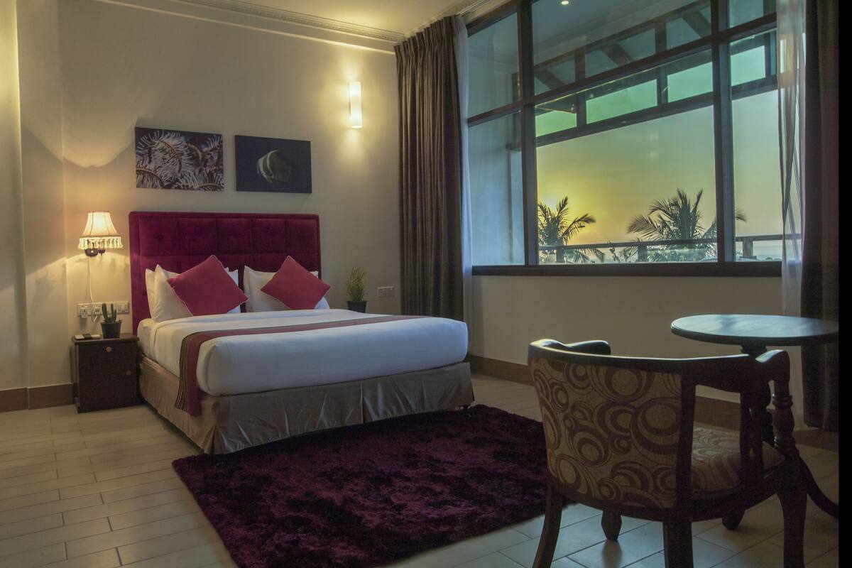 A hotel room with a bed, a nightstand, a lamp, a chair, a small table, and a window with a view of palm trees.
