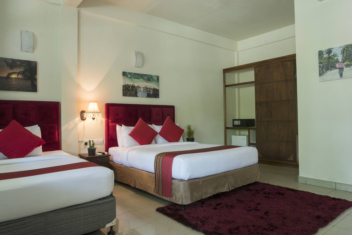 A hotel room with two beds, a large mirror, a red headboard, a nightstand with a lamp, and a red rug.