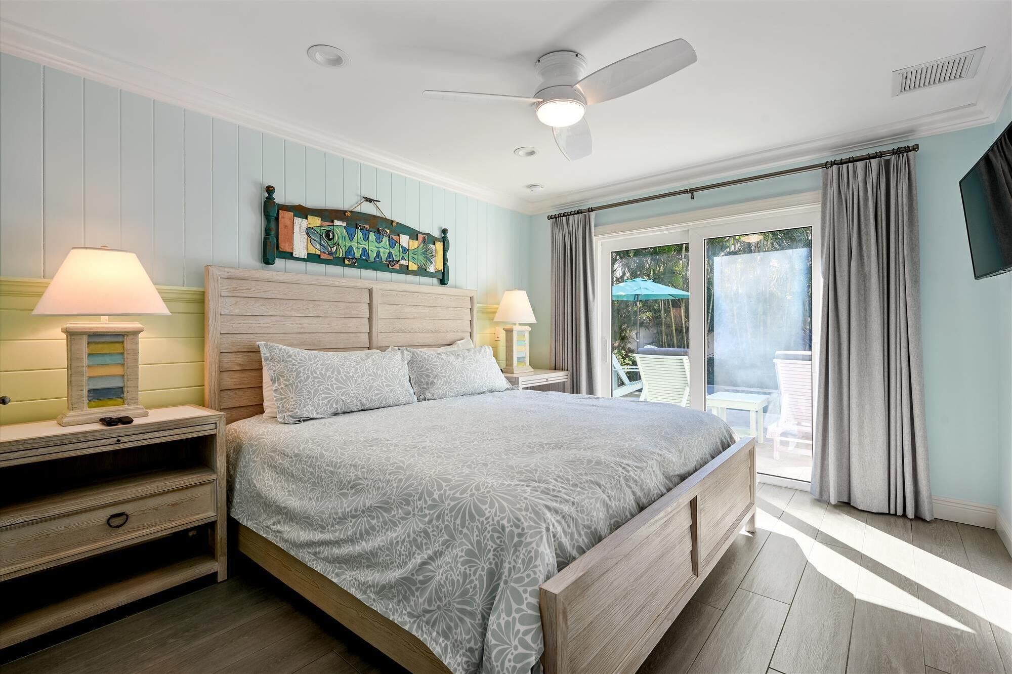A bedroom with a bed, two bedside tables with lamps, a ceiling fan, and a view to an outdoor area.