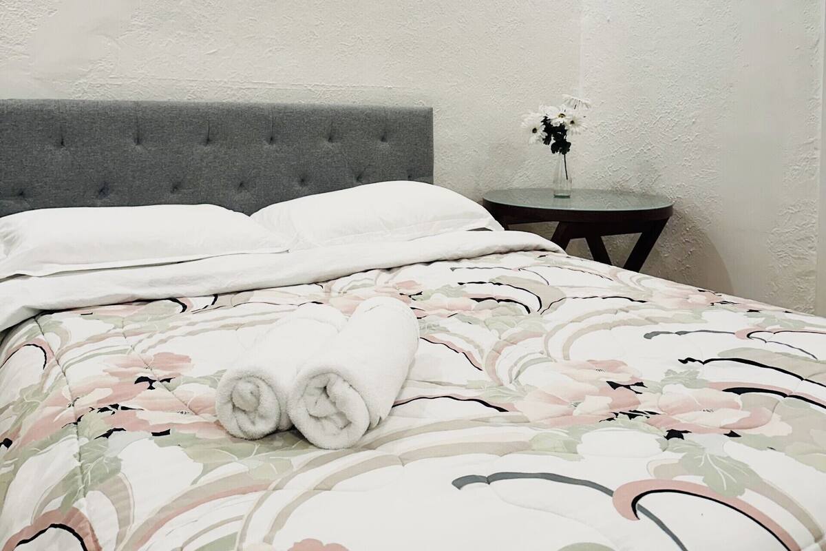 Un lit bien fait, agrémenté d’une couette à motifs floraux, d’une tête de lit rembourrée de couleur grise et d’une petite table de chevet sur laquelle est posé un vase de fleurs.
