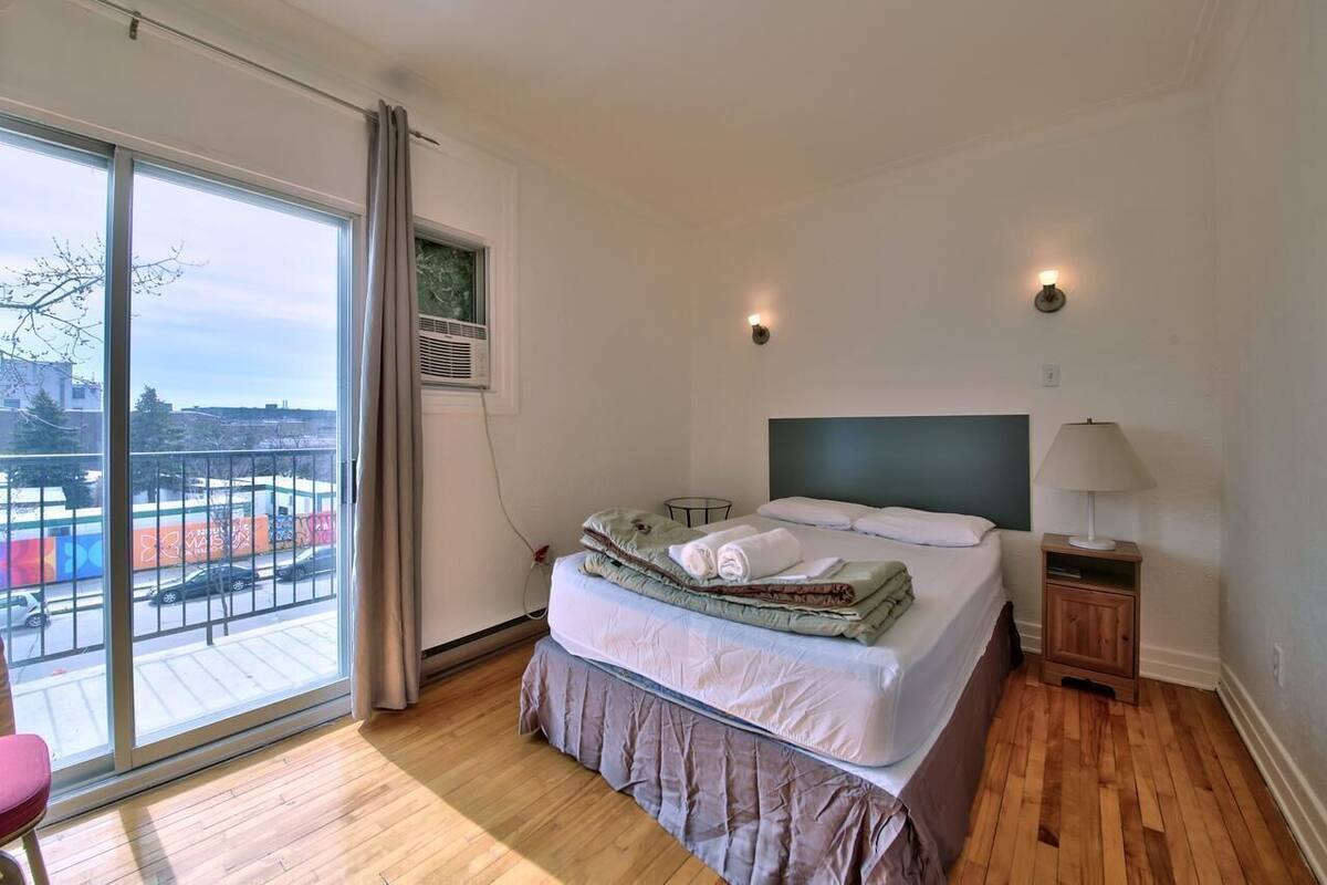Une chambre avec une grande fenêtre, un lit, une table de chevet et une vue sur la ville.