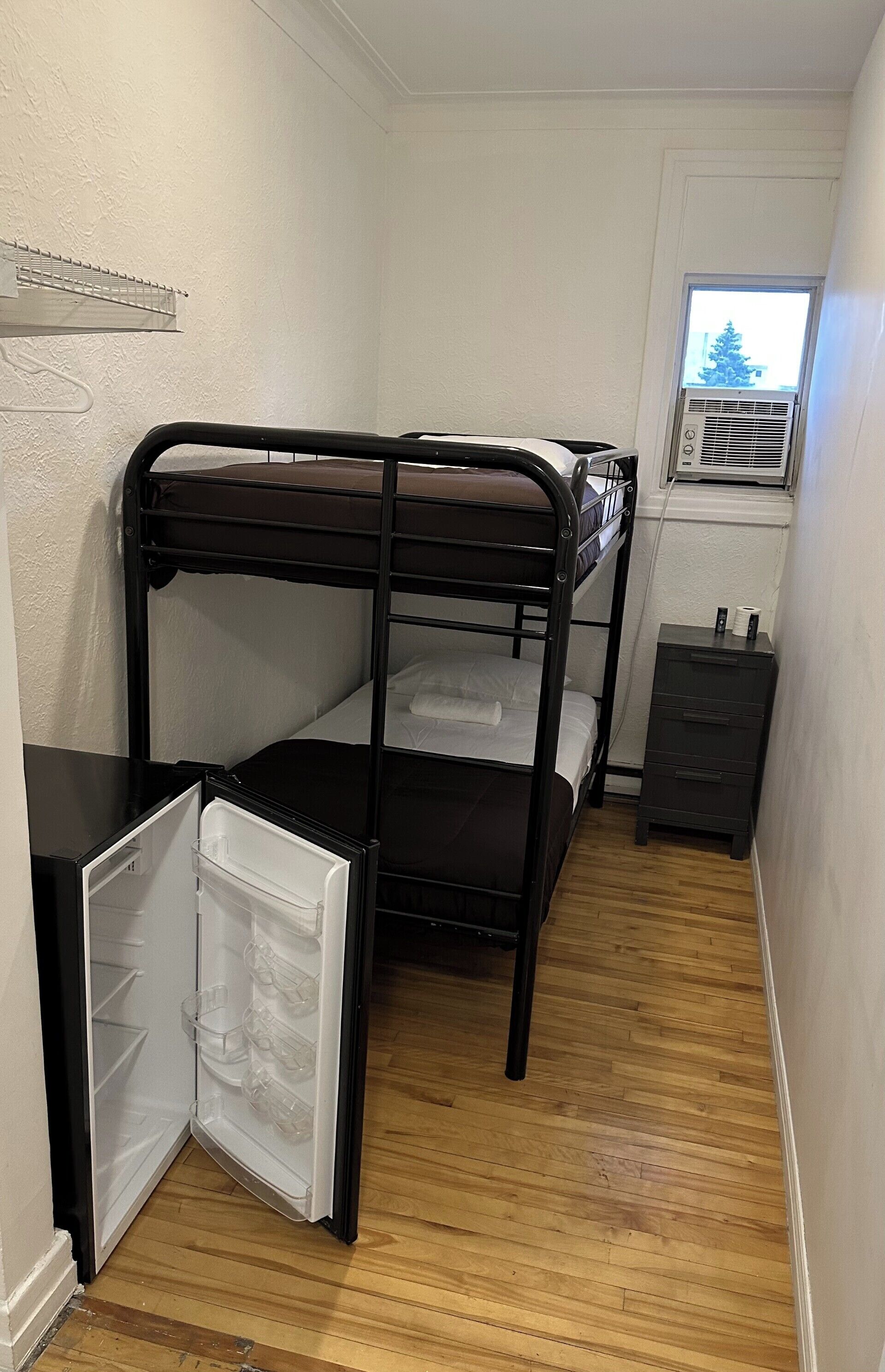 A bunk bed, a small dresser, and a refrigerator in a narrow room with wooden flooring.