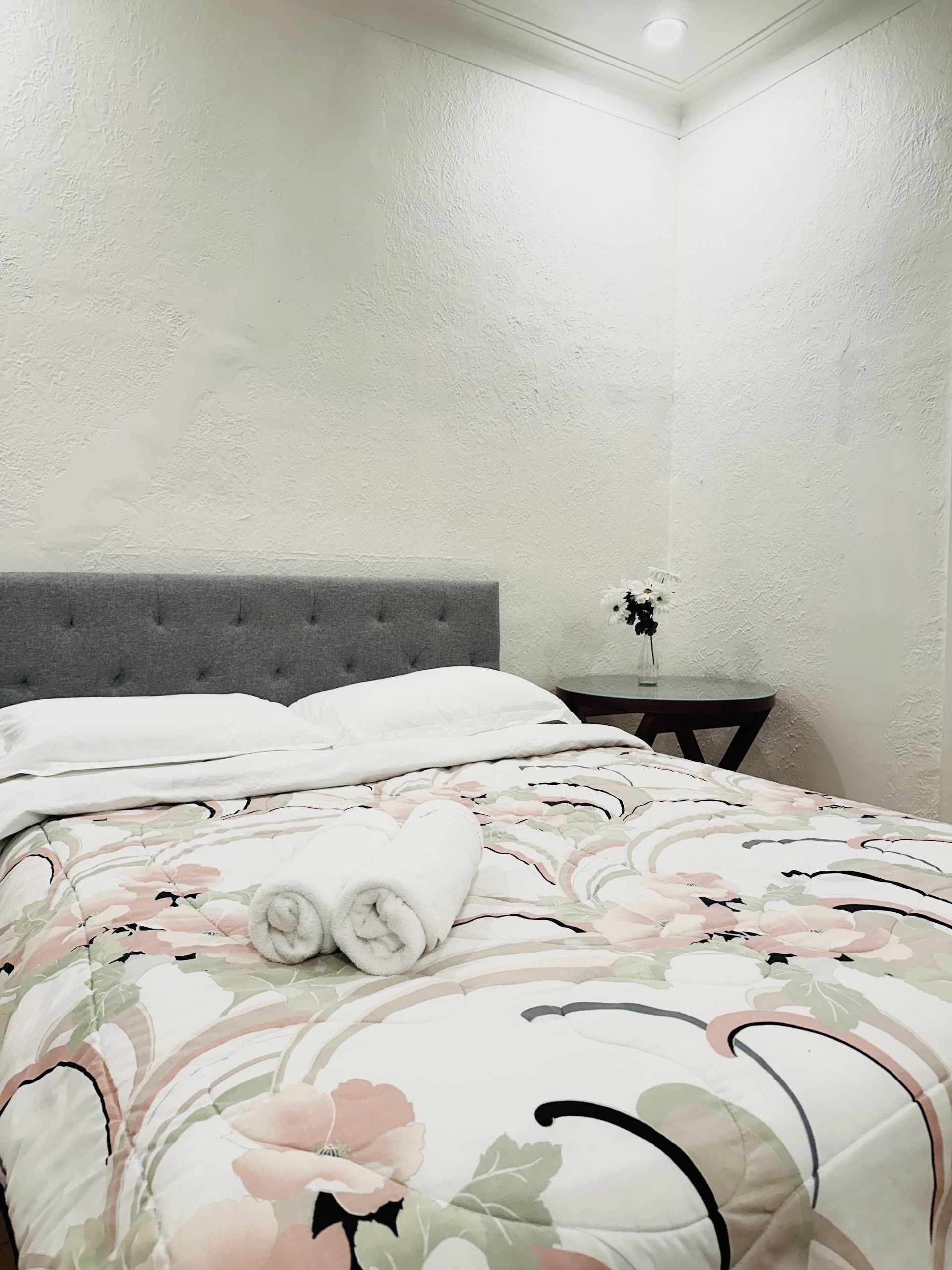 Una cama bien tendida con una colcha floral, un cabecero tapizado en gris y una mesita de noche con un florero.