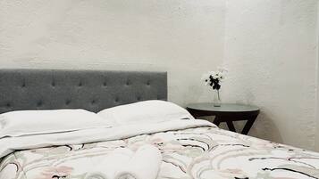 Una cama bien tendida con una colcha floral, un cabecero tapizado en gris y una mesita de noche con un florero.