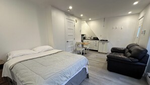 Un dormitorio con cama, una silla, un sofá y una kitchenette al fondo.