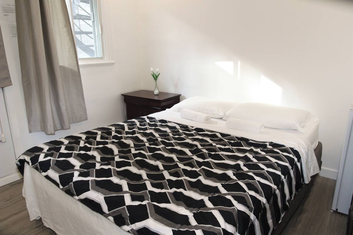 Une chambre à coucher avec un lit, une table de chevet, un vase de fleurs et une fenêtre avec des rideaux.
