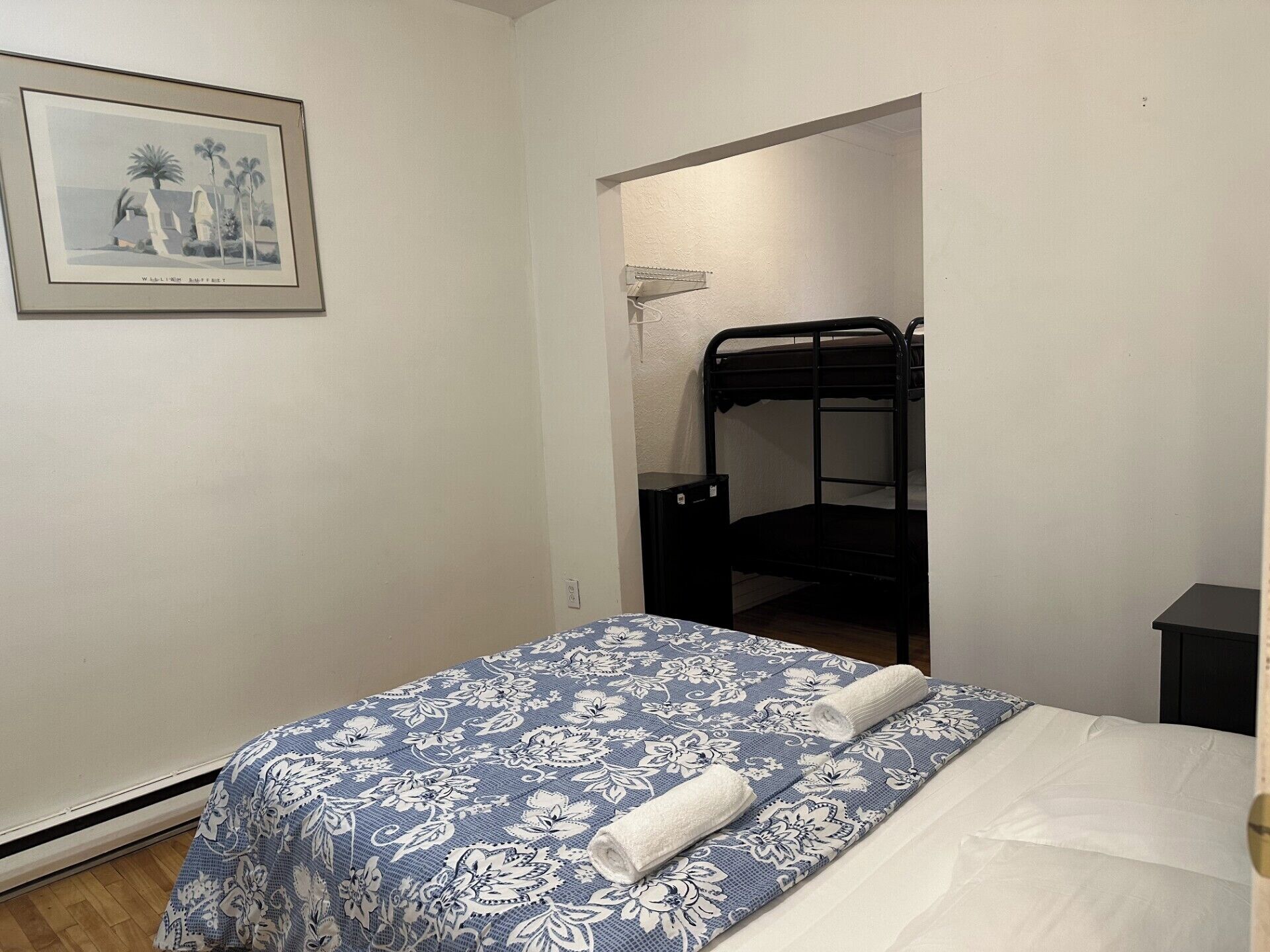 A bedroom with a bunk bed, a bed with a floral bedspread, a framed picture on the wall, and a small bedside table.