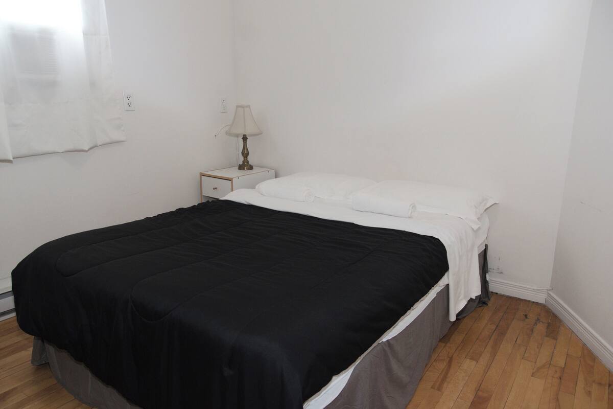 Une chambre à coucher avec une couvre-lit noire, des oreillers blancs, une table de chevet avec une lampe et un plancher en bois.