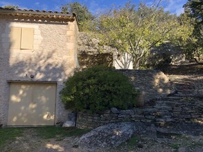 Un edificio rústico de piedra con una puerta de garaje, rodeado de follaje y un muro de piedra.