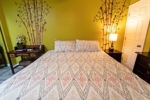 A bedroom with a patterned bedspread, a bedside table with a lamp, a dresser, and a wall decoration of bamboo branches.