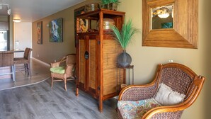 A living room with a wooden cabinet, wicker chairs, a patterned sofa, and a large mirror on the wall. - Hale Kamaole 127 Ground floor condo, easy access to best beach on Maui! (Kihei)