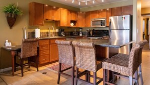 A modern kitchen with wooden cabinets, a stainless steel refrigerator, and a central island with bar stools. - Hale Kamaole 127 Ground floor condo, easy access to best beach on Maui! (Kihei)