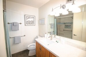 A bathroom with a white sink, wooden cabinet, and a large mirror. There is a towel rack with two towels and a framed picture on the wall. - #128 Ground floor condo, easy access to best beach on Maui! (Kihei)