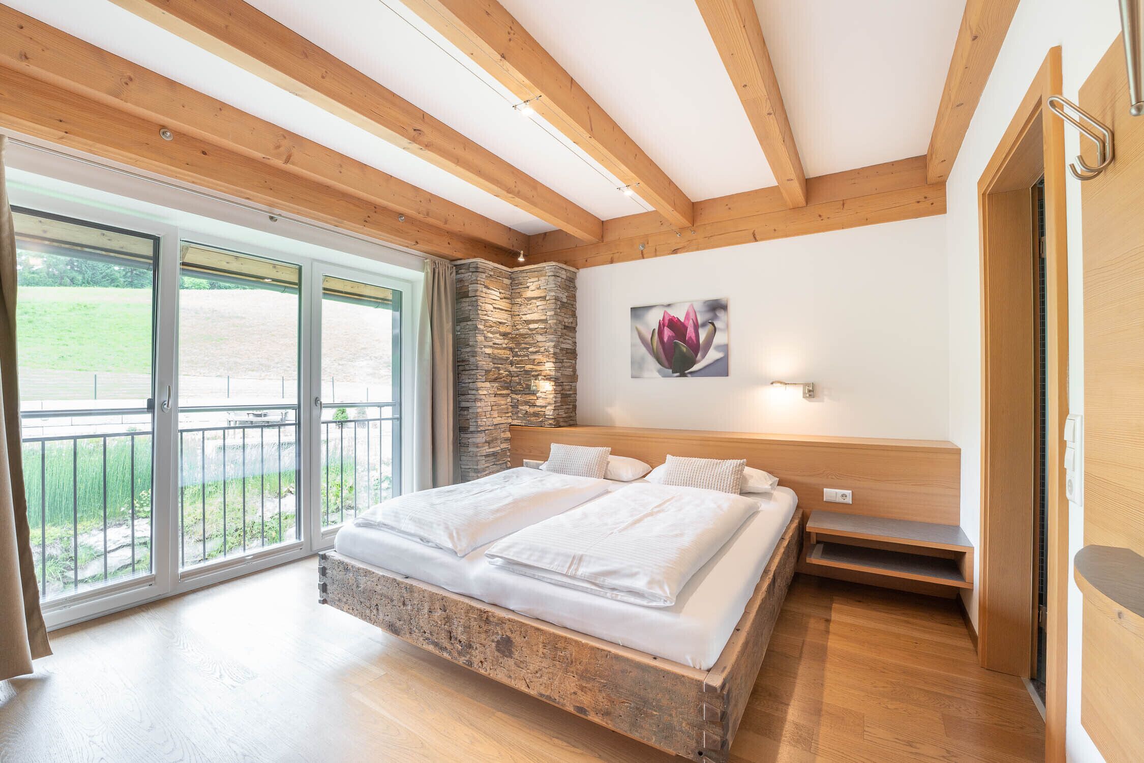 Une chambre dotée d’un grand lit, de poutres en bois, d’un mur en pierre et d’un balcon offrant une vue sur la verdure.
