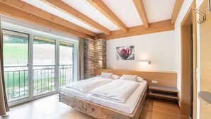 Quarto com cama grande, vigas de madeira, parede de pedra e varanda com vista para o verde.