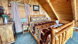 Um quarto de sótão aconchegante, com teto inclinado de madeira, uma cama com colcha estampada, uma pequena mesa de cabeceira de madeira, uma cadeira e uma janela com cortinas.