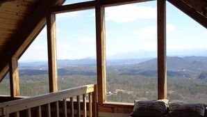 Quarto com grande janela que oferece vista para uma paisagem montanhosa. Há um corrimão de madeira e uma cama com roupa de cama listrada.