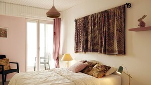 A bedroom with a bed, a chair, a lamp, and a window with curtains. - Flat for 6 on the main beach, St Gilles (Saint-Gilles-Croix-de-Vie)